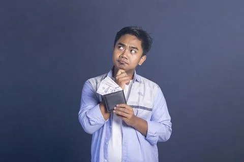 A thoughtful young man looking up while holding a black wallet filled with .. Stock-Fotos