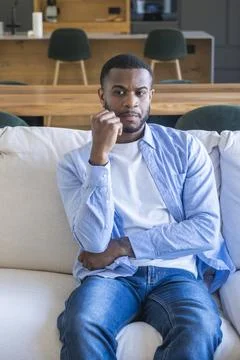 A thoughtful young man sits on a sofa at home, arms crossed, looking pensiv.. Stock Photos