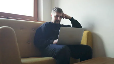 Thoughtful young man working on laptop on yellow sofa at home Stock Footage 337897105