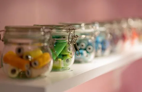 Threads spools in glass jars closeup on white shelf in pink wallpaper room Stock Photos