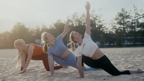 Three athletic beautiful girls doing yoga exercises on a sandy beach at dawn on Stock Footage 252138818
