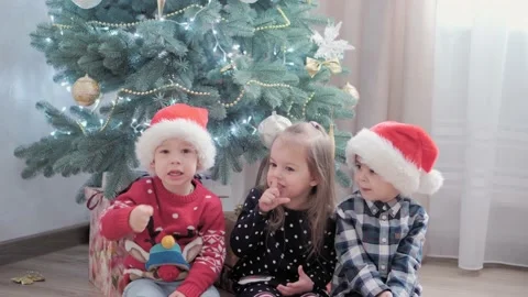 Three authentic happy Joy children wave hands hello sit by Christmas tree play Stock Footage 153882030