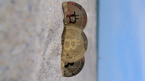 Three Bitcoin BTC coins closeup Bitcoin BTC coin half buried sand beach Vertical Stock Footage 296120825