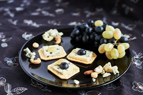 Three blue cheese crackers with grapes are on a black plate. Stock Photos