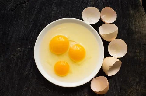 Three broken raw eggs on a white plate stand on a dark kitchen table Stock Photos