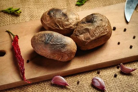 Three brown potatoes are on a wooden cutting board with garlic cloves, dried  Stock Photos