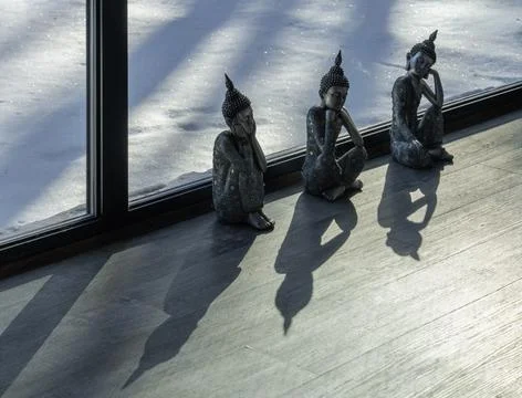 Three Buddha figurines by window with long shadows on wooden floor Stockfoto's