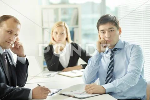 Three busy colleagues sitting at table and looking at camera Stock Photos