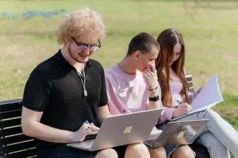 Three cheerful students study and work together outdoors on a sunny day, using Stock Photos