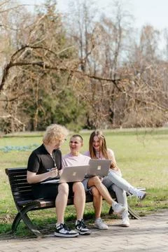 Three cheerful students study and work together outdoors on a sunny day, using 스톡 사진