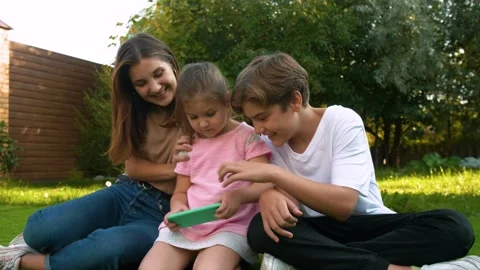 Three children are sitting on the grass and playing on a smartphone. Stock Footage 142500064
