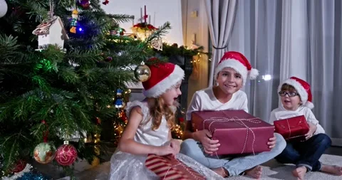 Three children in Santa hats pose with gifts near Christmas tree Video stock 294692452