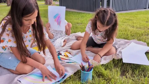 Three children sit on a blanket in a park. They draw on papers with crayons. Two Stock Footage 341490831