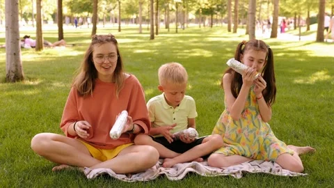 Three children sit in the park on the grass in summer and eat corn. Stock Footage 299904036