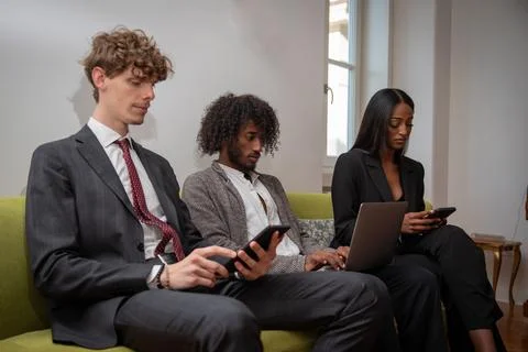 Three colleagues use their tech devices while sitting on the sofa at their .. 库存照片