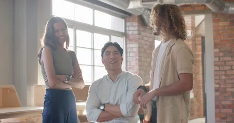 Three coworkers standing and talking in loft studio, seated man on stool Stock Footage 339983518