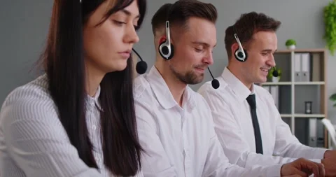 Three customer support agents in headsets working on laptops at desks indoors Stock Footage 344768032