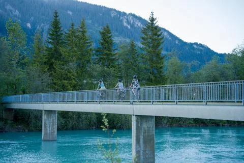 Three Cyclists Riding Across River Bridge With Mountain Forest Backdrop at Du Stock Photos