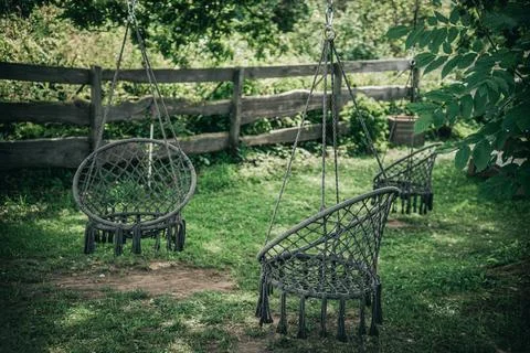 Three dark wicker hanging chairs are hung under deciduous. Stock Photos