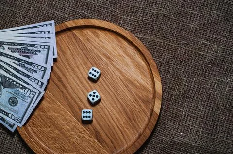 Three dice laying over a pile of money on a wooden plate Stock Photos