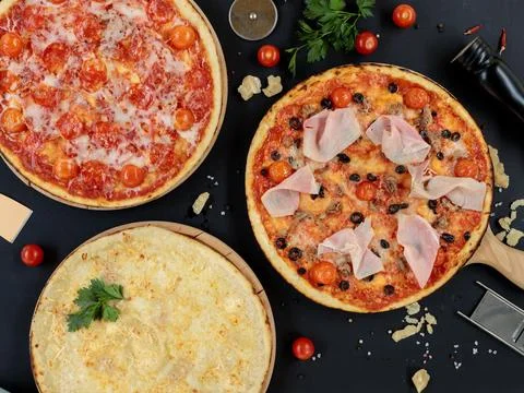 Three different pizzas on a black background with cooking ingredients. Top view Stock Photos