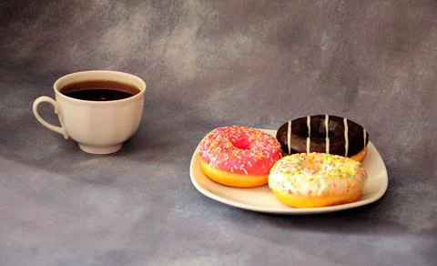 Three donuts in multicolored glaze on a white ceramic plate and a cup of blac Stock Photos