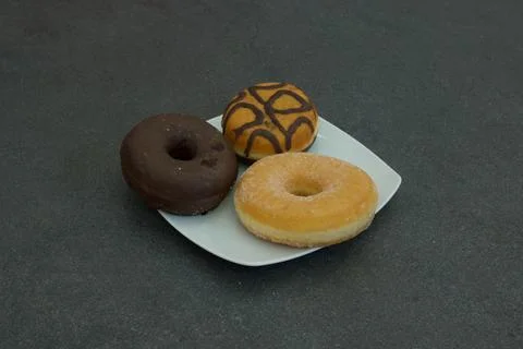 Three donuts on a plate, one of which is chocolate and the other two are glaz Stock Photos