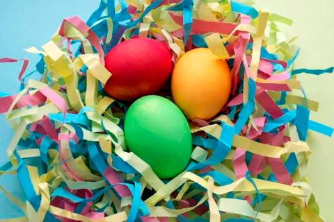 Three dyed Easter eggs in a colorful nest on mixed blue and yellow background. Stock Photos