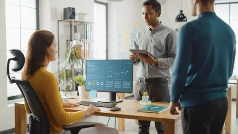 Three Economic Analytics Work on Desktop Computer, They Discuss Statistics Data Video stock 112601786