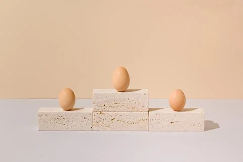 Three eggs standing on a travertine marble podium on a beige and gray background Stock Photos