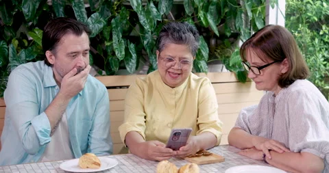 Three elderly friends laughing while looking at smartphone together at cafe, Stock Footage 308060475