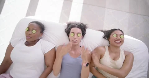 Three female friends lying on bed, center woman placing cucumber slices for home Stock Footage 335895202