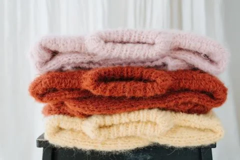Three folded multi-colored sweaters lie on a stool in front of a curtain. Stock Photos