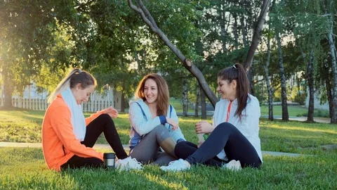 Three friends are sitting on the grass after a morning jog, they laugh and talk. 스톡 동영상 119047499