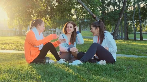 Three friends are sitting on the grass after a morning jog, they laugh and talk. Stock Footage 119047605