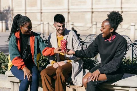 Three friends exchanging presents while sitting on a bench on a square Stock Photos