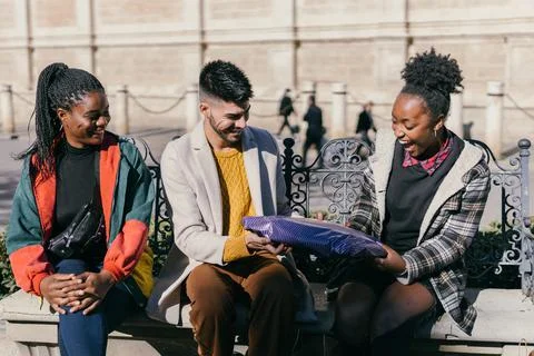 Three friends exchanging presents while sitting on a bench outdoors Stock Photos