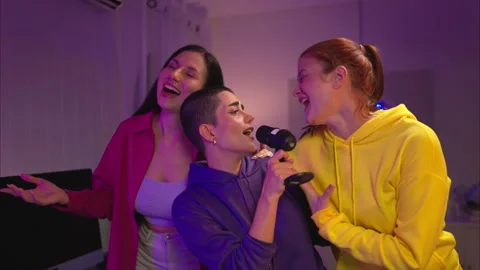 Three friends singing together into a microphone in a fun, neon-lit room Video stock 326223382