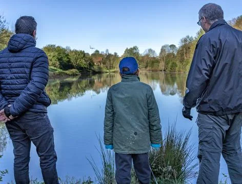 Three generations of men standing at lake shore looking at water together. Stock-Fotos