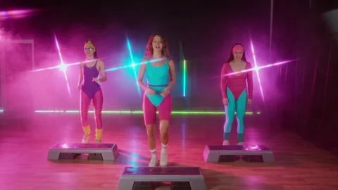 Three Girls Beginning Retro Step Aerobics Routine in Neon Lit Exercise Hall Stock Footage 328367179