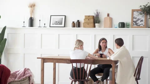 Three girls sit at a table and on chairs in a large room at home and talk Stock Footage 131847891