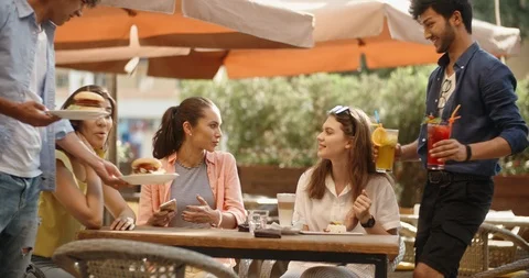 Three girls sitting at table in cafe chatting, then two guys approach with food Video stock 94605310