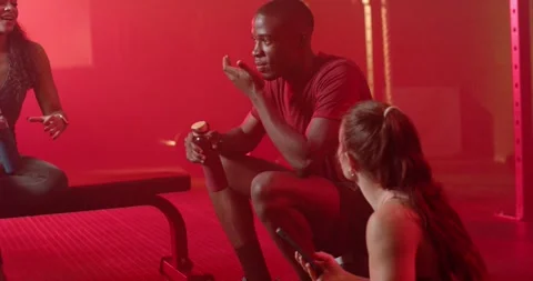 Three gym mates sitting on bench in red-lit gym, woman showing smartphone, man Stock Footage 339801669