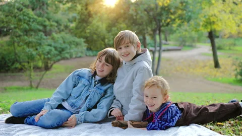 Three Happy Children Sitting Together on Blanket in Park During Golden Hour. Видео 338139663