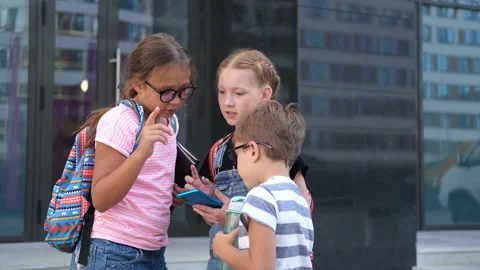 Three kids with backpack stand and talk together and play phone Stock Footage 157782874