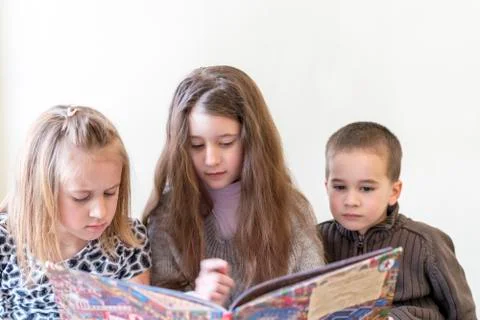 Three kids having fun reading a book. Two sisters and a brother. Light backgr Stock Photos