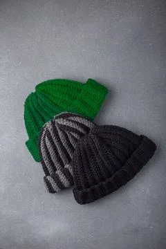 Three knitted woolen beanie caps or hats, black, green and striped on grey Stock Photos