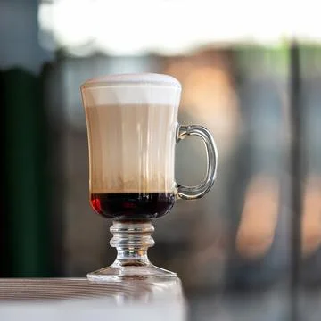 Three-layer coffee with foam in tall glass cup with handle on table against of Stock Photos