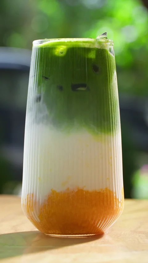 Three-layered mango-milk-matcha drink in a glass on a greenery background Stock Footage 314463103