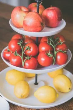Three-level plate with apples, tomatoes, and lemons on it in the kitchen Stock Photos